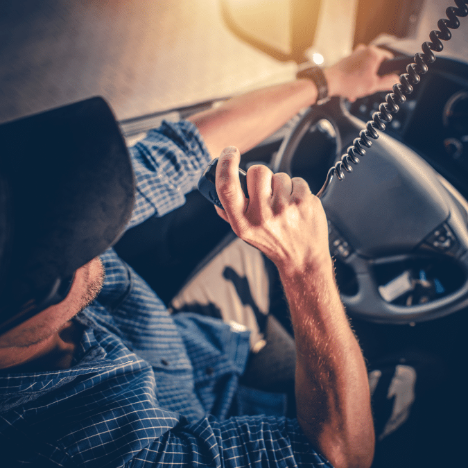 A person wearing a cap and blue plaid shirt drives a vehicle while speaking into a CB radio, with one hand on the steering wheel and sunlight streaming through the window.