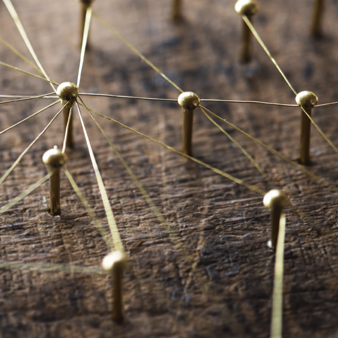 Close-up of brass pins connected by thin golden threads on a wooden surface, forming an abstract network or web pattern.