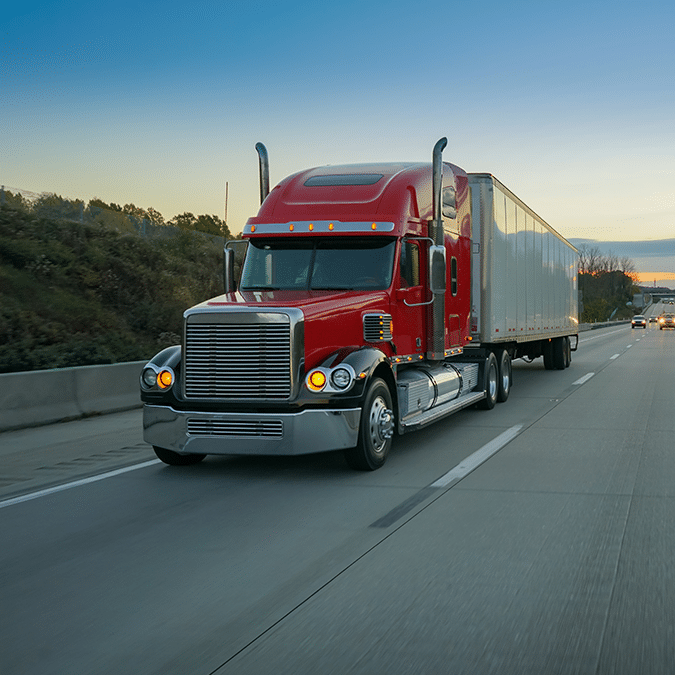 A red semi-truck with a large white trailer drives down a highway at dusk, passing green trees and grassy areas along the roadside under a clear blue sky.