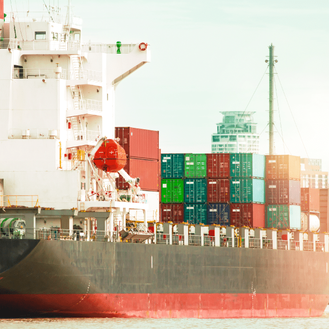 A cargo ship docked at a port, loaded with colorful shipping containers, with city buildings visible in the background.