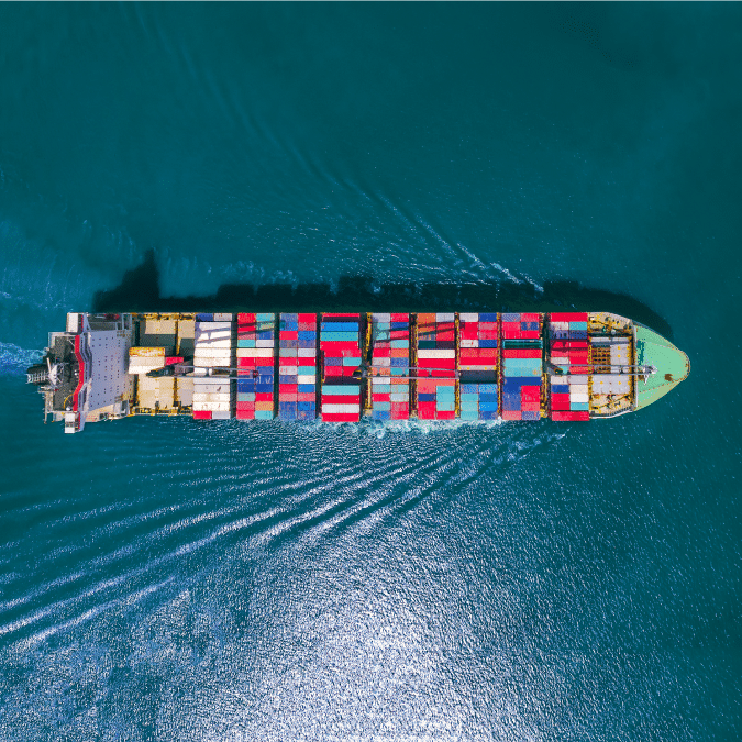 Aerial view of a large cargo ship carrying colorful shipping containers, sailing through deep blue ocean water and creating ripples behind it.