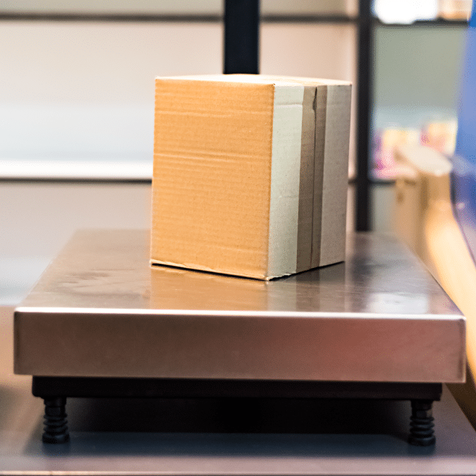 A small cardboard box sits on a metal platform scale, likely in a shipping or postal setting, ready to be weighed.