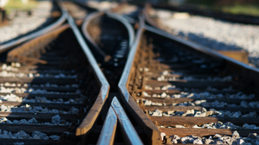 Close-up view of intersecting railway tracks with sunlight casting shadows, gravel surrounding the rails, and green scenery blurred in the background.