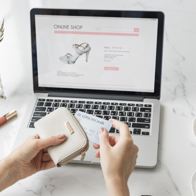 A person holds a beige wallet and a credit card while shopping for shoes online on a laptop. The laptop screen displays a sandal with reviews and a purchase option.