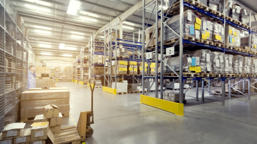 A spacious warehouse with tall shelves stacked with boxes and packages, a pallet jack, and organized aisles under a high ceiling with bright lighting.