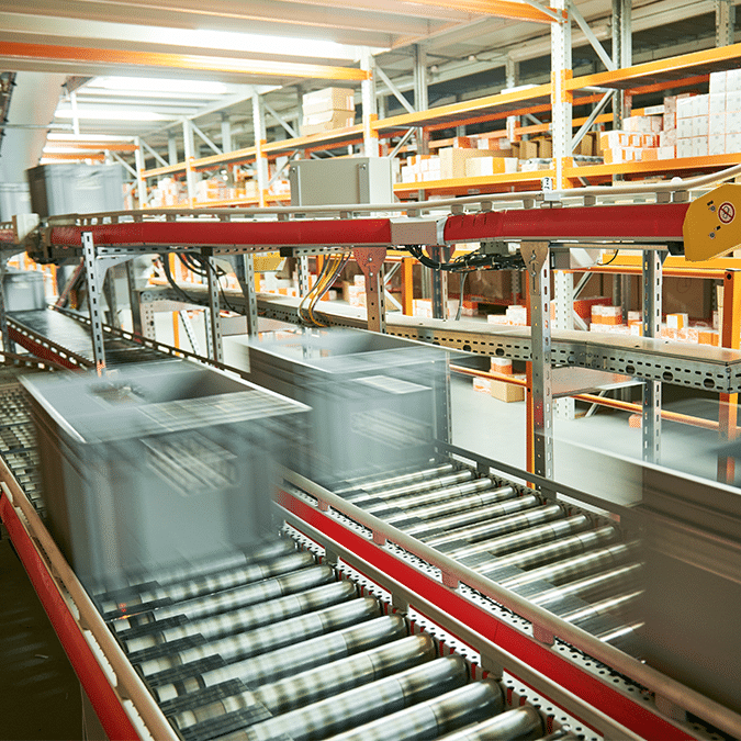 A warehouse conveyor system, designed with automation system engineering, transports large boxes rapidly across metal rollers, with shelves of orange and white packages in the background. The movement of the boxes appears blurred.