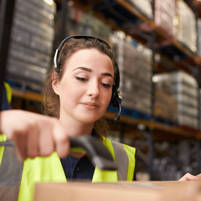 A warehouse worker wearing a headset and high-visibility vest scans a package with a barcode scanner in a storage facility with shelves full of boxes in the background.