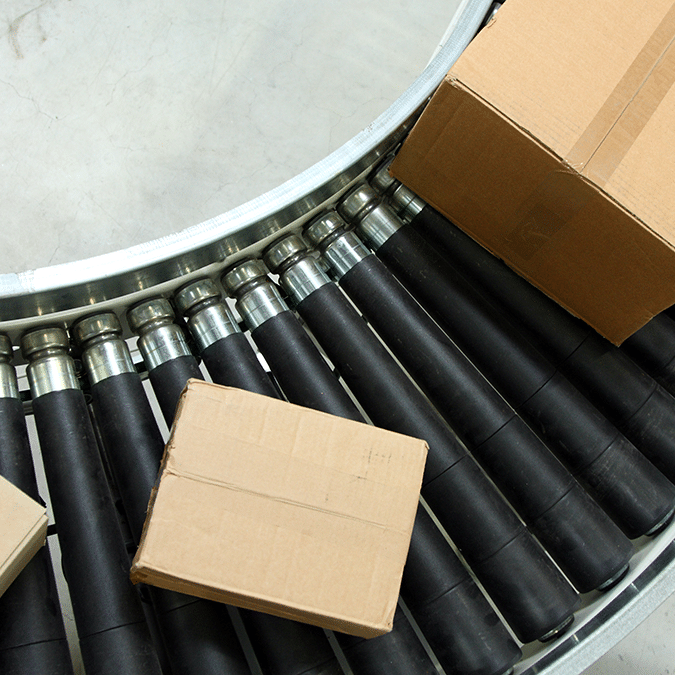 Three cardboard boxes on a curved, black roller conveyor belt, used for moving packages in a warehouse or shipping facility. The surface underneath is a smooth light gray.