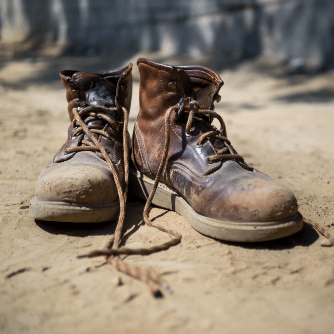 A pair of worn, brown leather work boots with untied laces are placed on sandy ground in sunlight, showing scuff marks and signs of heavy use.