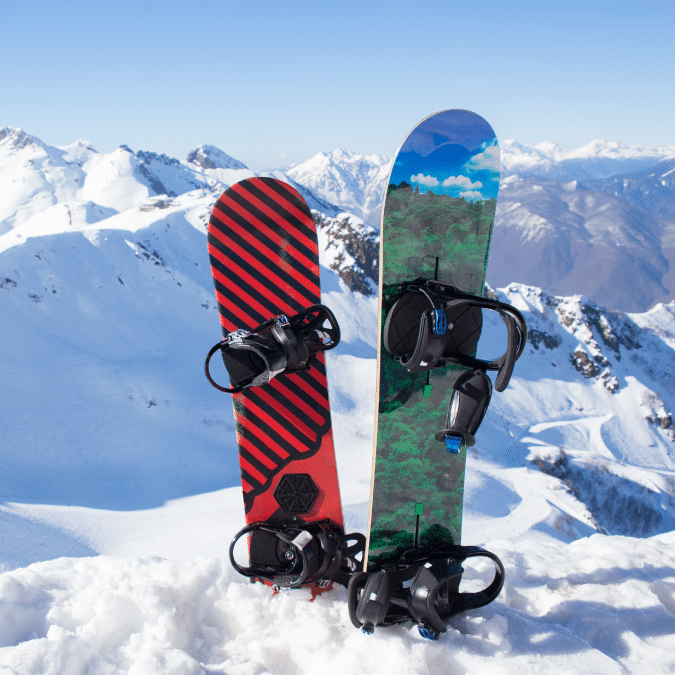 Two snowboards standing upright in snow on a mountain, one with red and black stripes and the other with a green and blue landscape design, with snowy peaks and a clear blue sky in the background.
