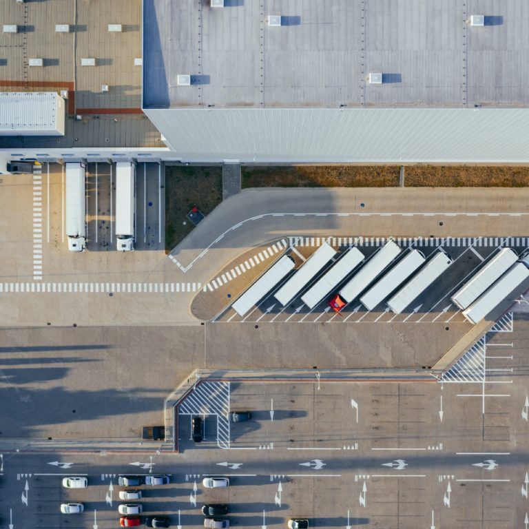 Aerial view of a large warehouse with multiple loading docks, several semi-trailers parked in a row, and a parking lot with cars in the lower section of the image.