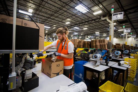 A warehouse worker in an orange safety vest packs items into a cardboard box at a workstation in a large, brightly lit industrial facility filled with equipment and stacks of boxes.