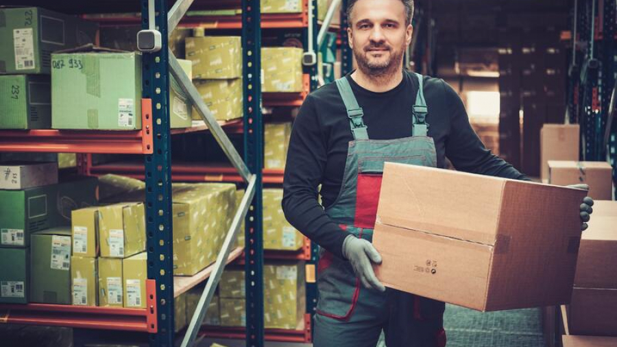 A man in work overalls and gloves stands in a warehouse, holding a large cardboard box. Shelves filled with various packed boxes are visible behind him. The environment is well-lit and organized.