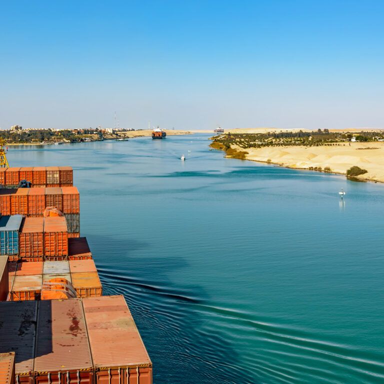 A large cargo ship with stacked shipping containers sails through the Suez Canal, bordered by sandy desert on the right and buildings in the distance under a clear blue sky.
