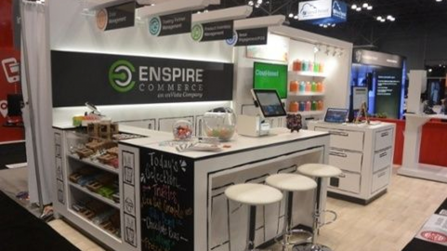 A trade show booth for Enspire Commerce with a countertop, white stools, computer screens, and organized shelves of snacks. Bright signs and colorful jars are displayed in a well-lit exhibition space.