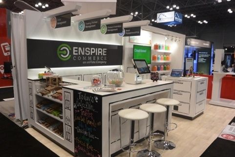 A trade show booth for Enspire Commerce with a countertop, white stools, computer screens, and organized shelves of snacks. Bright signs and colorful jars are displayed in a well-lit exhibition space.
