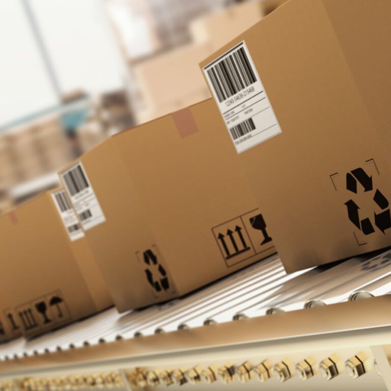 Several cardboard boxes with shipping labels and handling symbols move along a conveyor belt in a warehouse or distribution center. The background is blurred, emphasizing the boxes in the foreground.