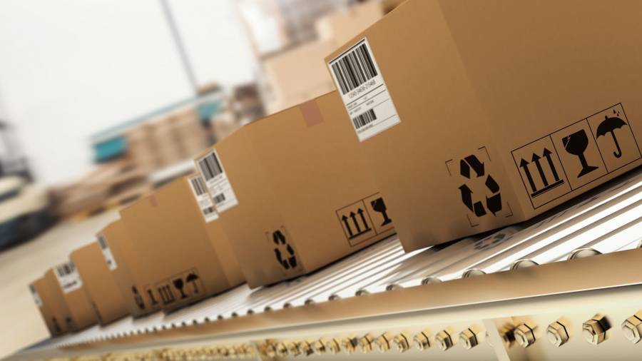 Several cardboard boxes with shipping labels and handling symbols move along a conveyor belt in a warehouse or distribution center. The background is blurred, emphasizing the boxes in the foreground.