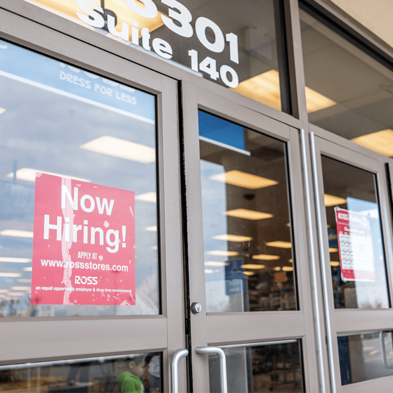 Glass doors of a retail store with a prominent red Now Hiring! sign displayed, reflecting the parking lot outside; store lights and aisles are visible through the glass.