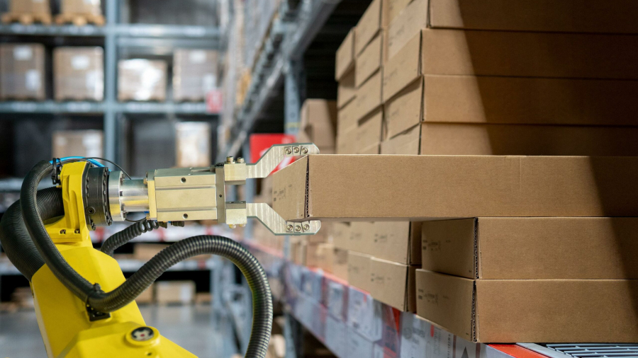 A robotic arm lifts a cardboard box from a shelf filled with similar boxes in a warehouse, illustrating automation in inventory management.