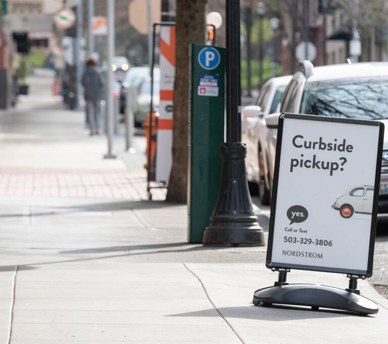 A sidewalk sign advertises curbside pickup for Nordstrom, featuring a graphic of a car and a phone number, with parked cars and people walking in the background on a city street.