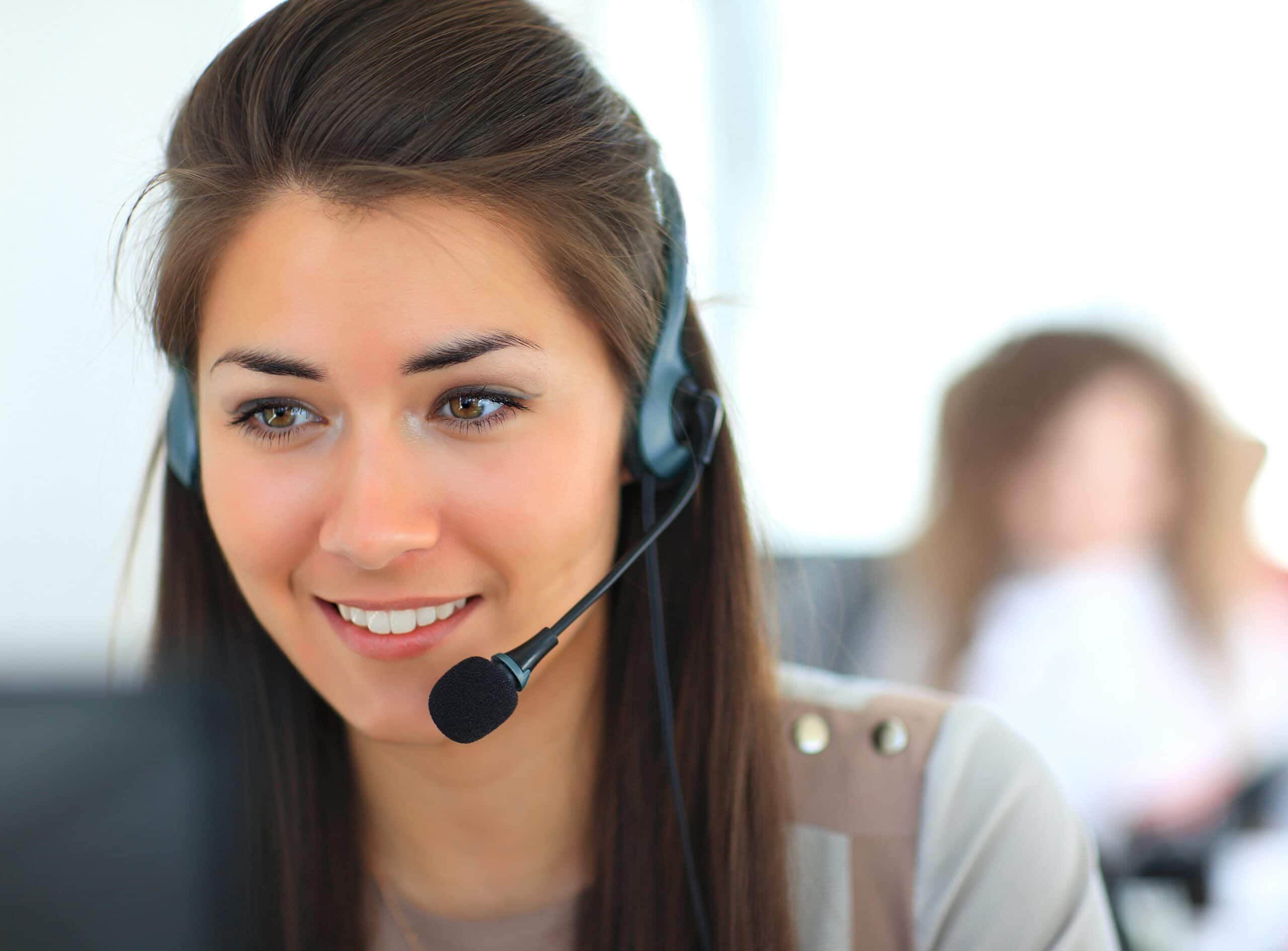 A woman wearing a headset with a microphone is smiling while looking at a computer screen, suggesting she is working in a customer service or call center environment. The background is blurred.