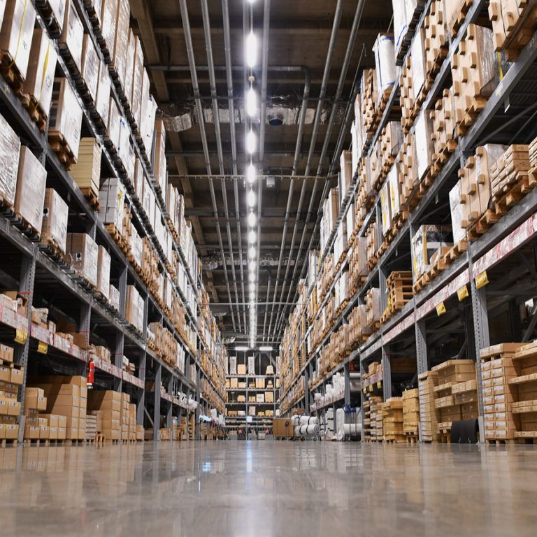 Wide-angle view of a warehouse aisle with tall shelves stacked with various cardboard boxes, illuminated by bright overhead lights and a shiny concrete floor—ideal for conducting a thorough warehouse capacity analysis.