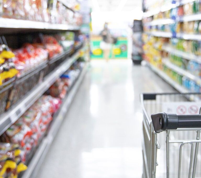 A shopping cart in a grocery store aisle, surrounded by shelves stocked with various packaged food items on both sides. The scene is brightly lit, and the shelves appear well-organized.