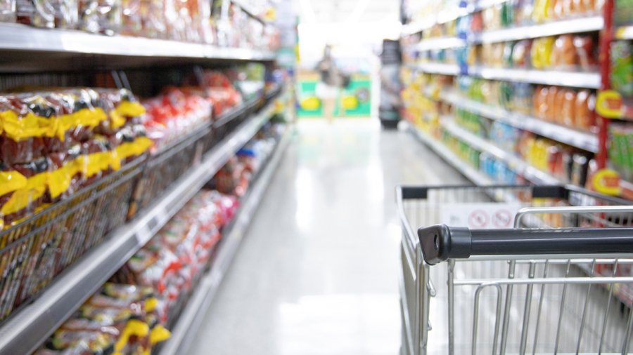 A shopping cart in a grocery store aisle, surrounded by shelves stocked with various packaged food items on both sides. The scene is brightly lit, and the shelves appear well-organized.