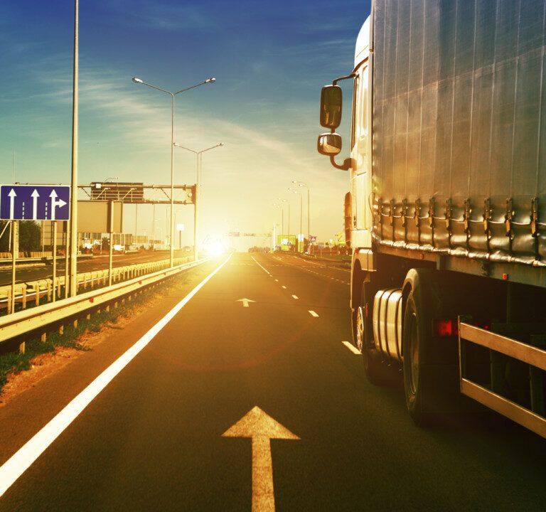A large truck drives down a highway at sunrise or sunset, with bright sunlight ahead, road signs on the left, and a clear sky overhead. The road features lane markings and guardrails.