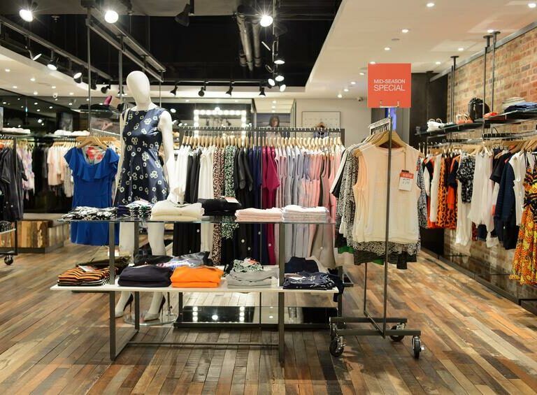 Clothing store interior with racks of colorful clothes, a mannequin in a blue dress, folded shirts on tables, a “Mid-Season Special” sign, wooden flooring, and bright ceiling lights reflects an efficient lean retail layout.