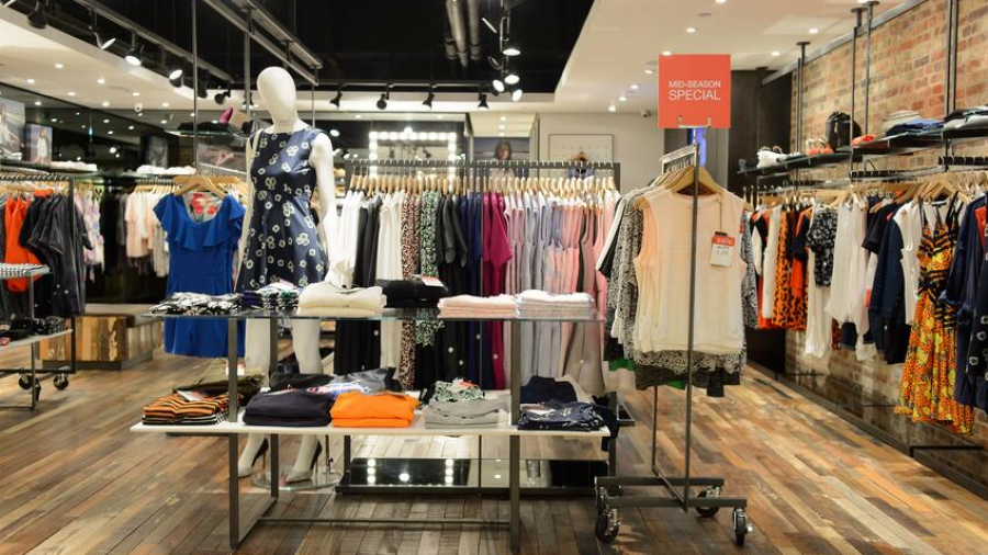 Clothing store interior with racks of colorful clothes, a mannequin in a blue dress, folded shirts on tables, a “Mid-Season Special” sign, wooden flooring, and bright ceiling lights reflects an efficient lean retail layout.