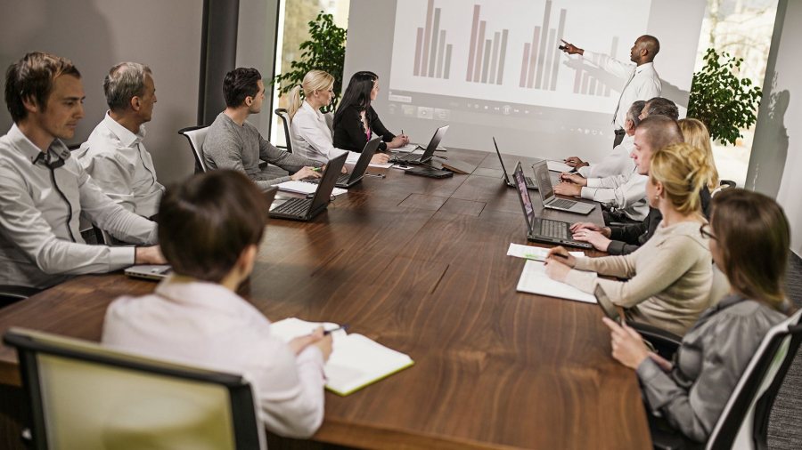 A group of people in business attire sit around a conference table with laptops and notepads, as a presenter stands near a screen displaying a bar graph during a Microsoft Dynamics AX upgrade meeting.