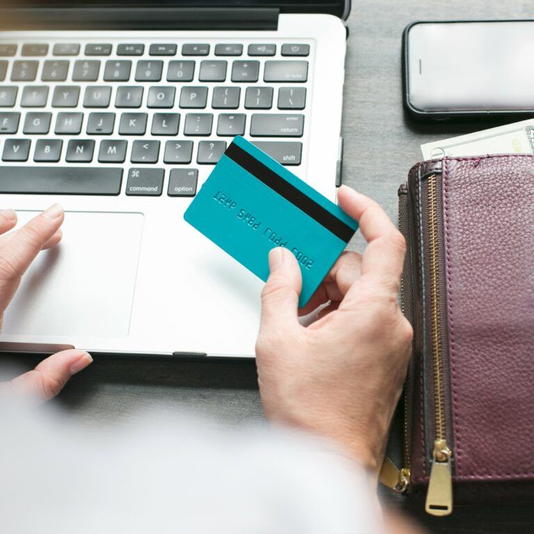 A person holding a blue credit card sits at a laptop, with a purple wallet, cash, coins, and a smartphone on the table nearby. The scene suggests online shopping or banking.