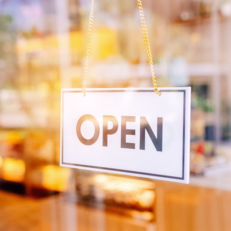 A white rectangular sign with the word “OPEN” in bold black letters hangs on a glass door, with a blurred interior of a shop or café visible in the background.