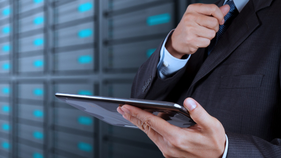 A person in a suit holds a tablet and adjusts their tie, standing in front of blurred server racks in a data center.