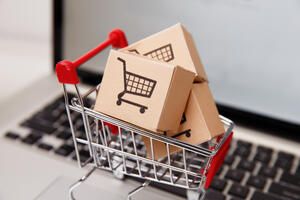 A small shopping cart filled with cardboard boxes, each featuring a shopping cart icon, sits on a laptop keyboard, representing online shopping or e-commerce.