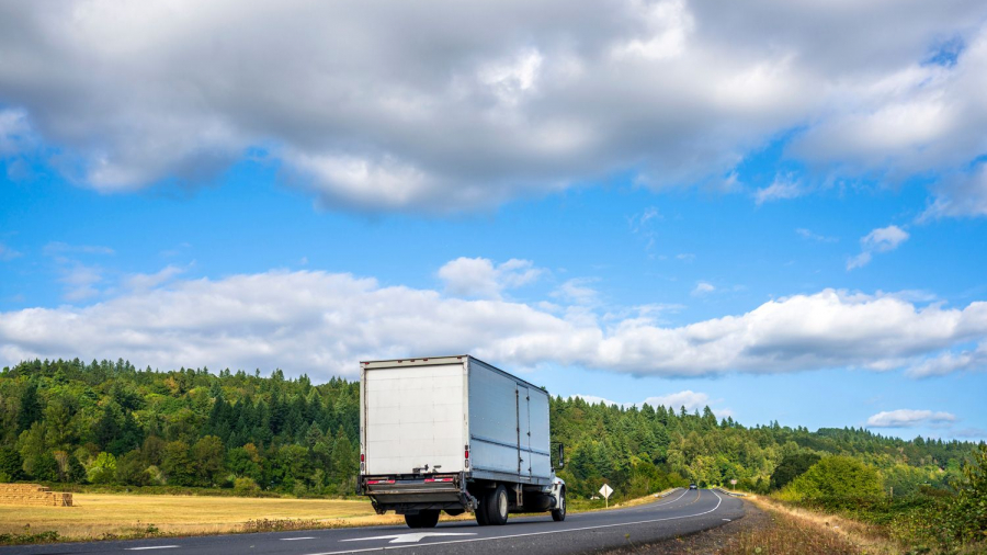 A white semi-truck drives down a curving rural road bordered by green trees and fields under a partly cloudy blue sky.