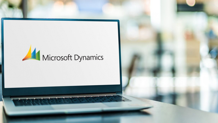 A laptop on a table displays the Microsoft Dynamics logo on its screen, with a blurred office environment in the background.