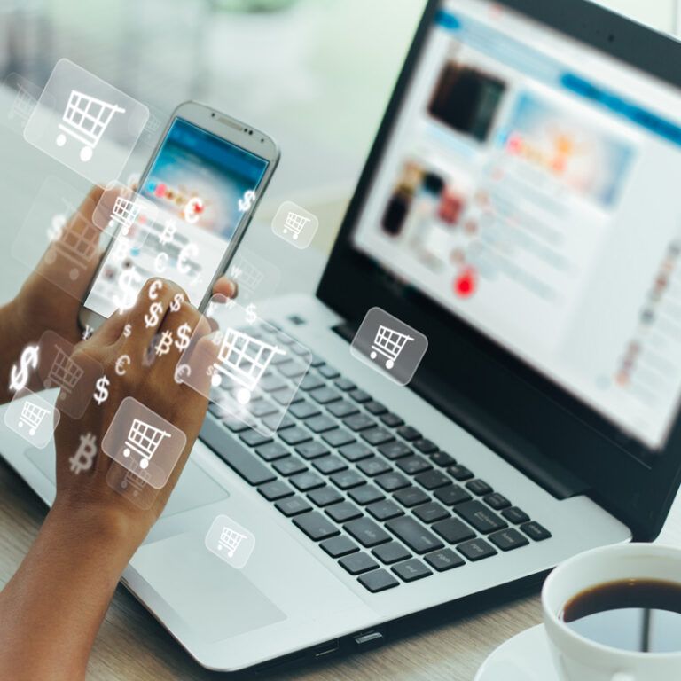 A person uses a smartphone and laptop simultaneously at a table with a coffee cup, with digital shopping cart, dollar, and currency symbols overlaying the image, representing online shopping or e-commerce.