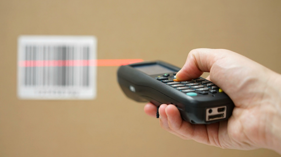 A hand holds a barcode scanner, using a red laser to scan a barcode on a cardboard box in the background.