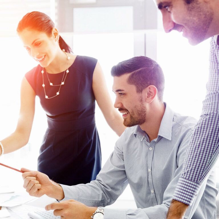 Three colleagues, two men and one woman, collaborate at a desk in a bright office, smiling and looking at a computer screen while discussing work. The woman is pointing at the monitor.