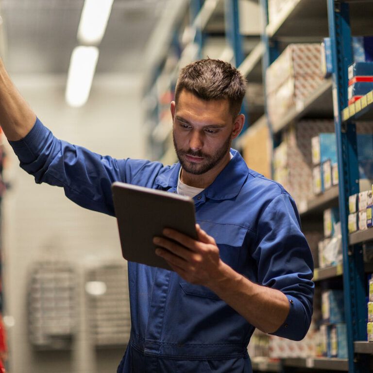 A man in a blue work uniform stands in a warehouse aisle, holding a tablet in one hand and reaching for a box on a shelf with the other. Shelves around him are filled with various small boxes and items.
