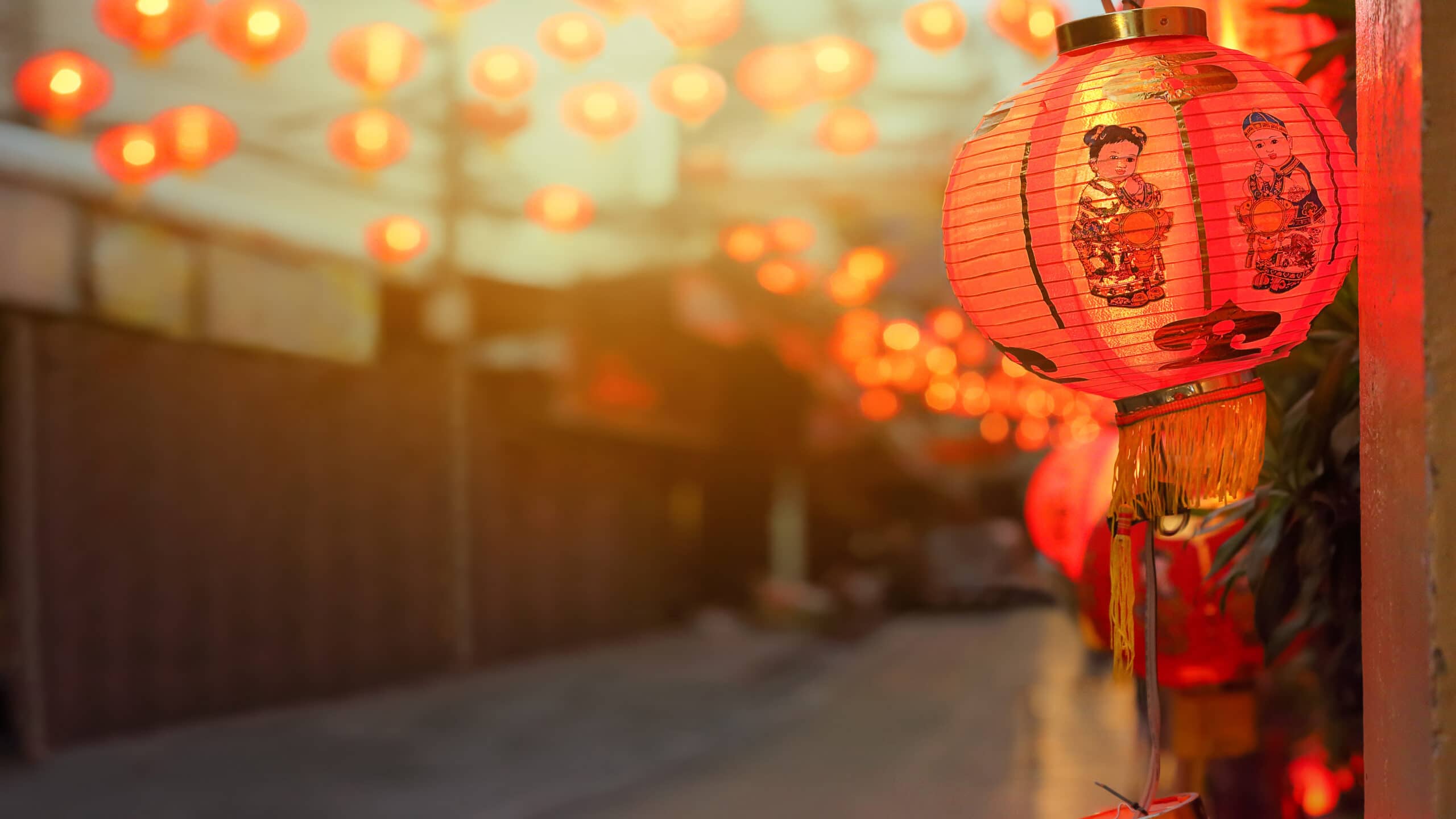 Red Chinese lanterns hang along a street at dusk, with one lantern in focus showing an illustration of a woman in traditional clothing. The background is softly blurred, filled with more glowing lanterns.