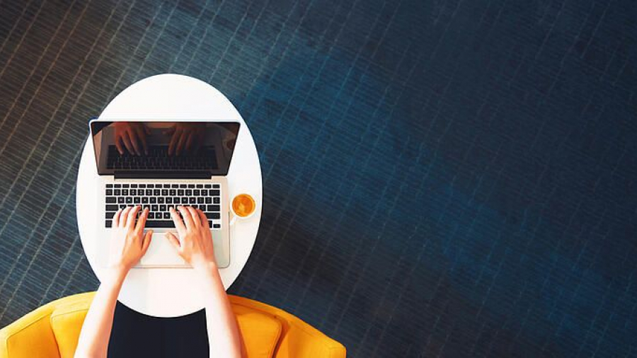A person types on a laptop at a white round table with a cup of coffee, seated in a yellow chair on a dark blue carpet. The image is viewed from above.