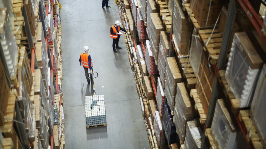 High-angle view of two workers in orange safety vests and white helmets moving and inspecting goods in a large warehouse filled with tall shelves stacked with boxes and pallets.