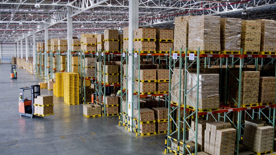 A spacious, well-lit warehouse with high shelves stacked with cardboard boxes, organized racks of goods in rows, and a worker operating a forklift—motivated by incentive based pay—on the left side of the image.