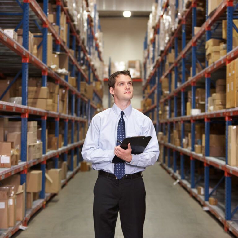 A man in business attire holds a clipboard while standing in the aisle of a warehouse, surrounded by tall shelves filled with cardboard boxes.