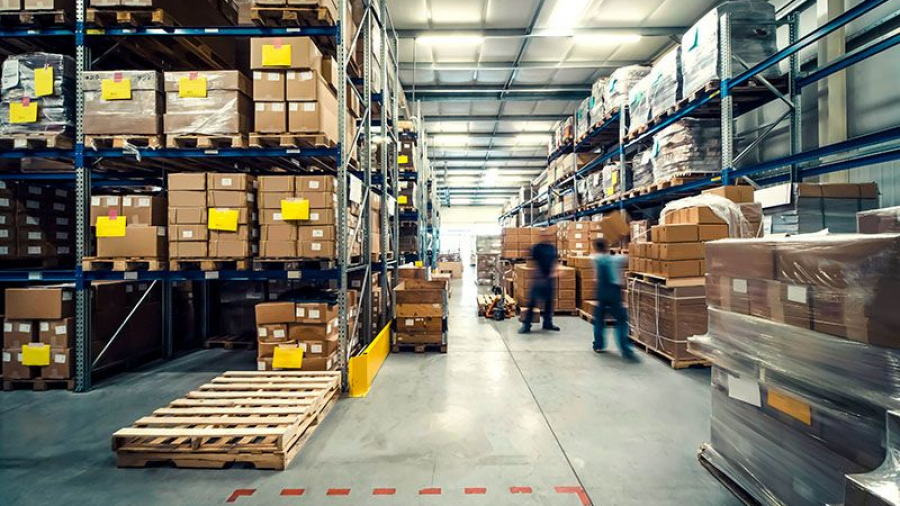 A spacious warehouse optimized for warehouse space utilization, with shelves stacked high with boxes and packages. Two workers move through the aisle, and pallets are scattered on the well-lit floor under overhead lighting.