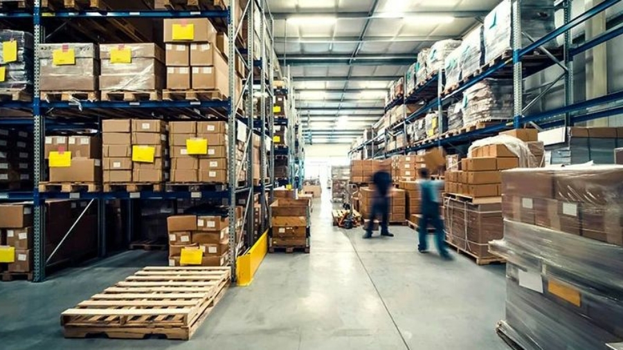 A warehouse with tall shelves stacked with boxes and pallets. Two blurred figures move along the aisle, driven by incentive based pay. The space is well-lit with overhead lights and organized for storage and distribution.